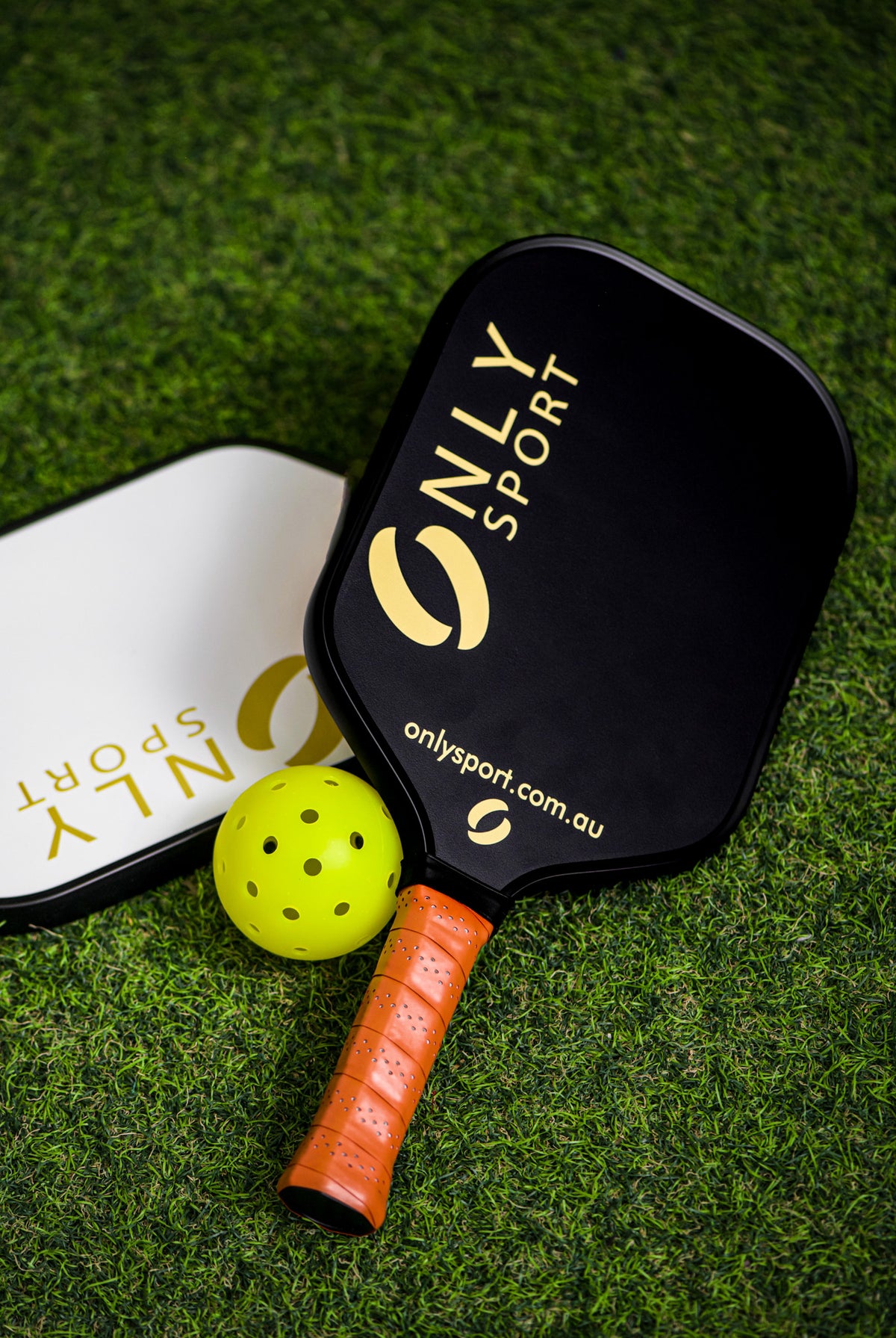 Displaying black paddle on top of white paddle on an angle next to a ball, laying on the artificial turf