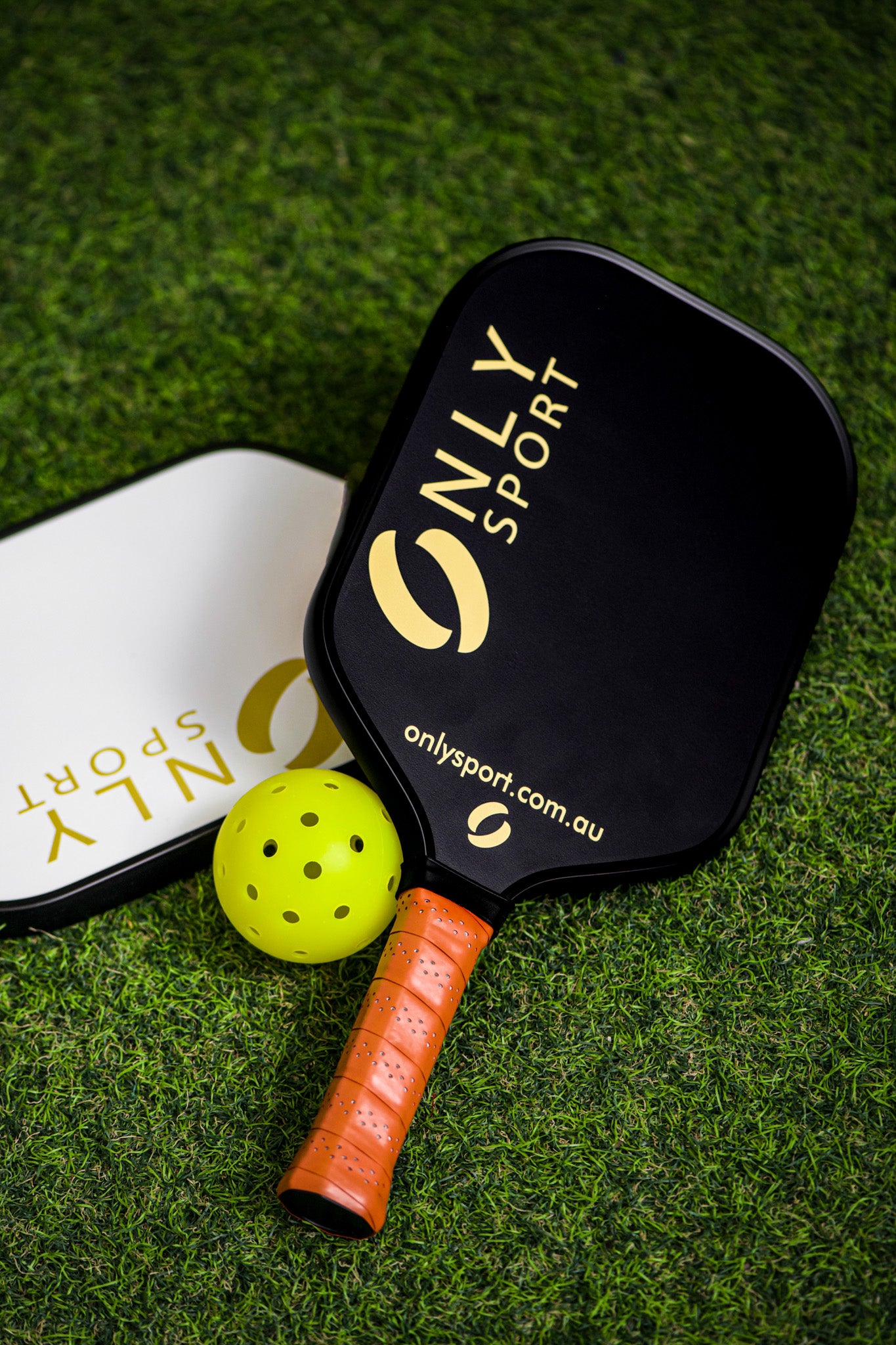 Displaying black paddle on top of white paddle on an angle next to a ball, laying on the artificial turf