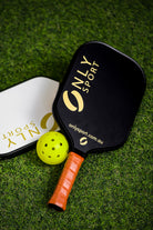 Displaying black paddle on top of white paddle on an angle next to a ball, laying on the artificial turf